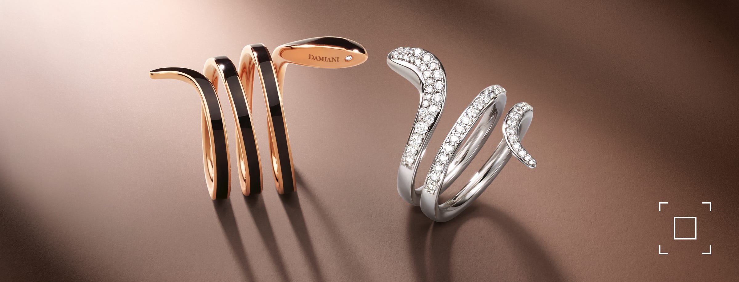 Anelli Damiani oro rosa con ceramica nera e oro bianco con pavè di diamanti bianchi su fondo marrone con ombra pronunciata. I due anelli a forma di serpente sembrano guardarsi, innamorati.